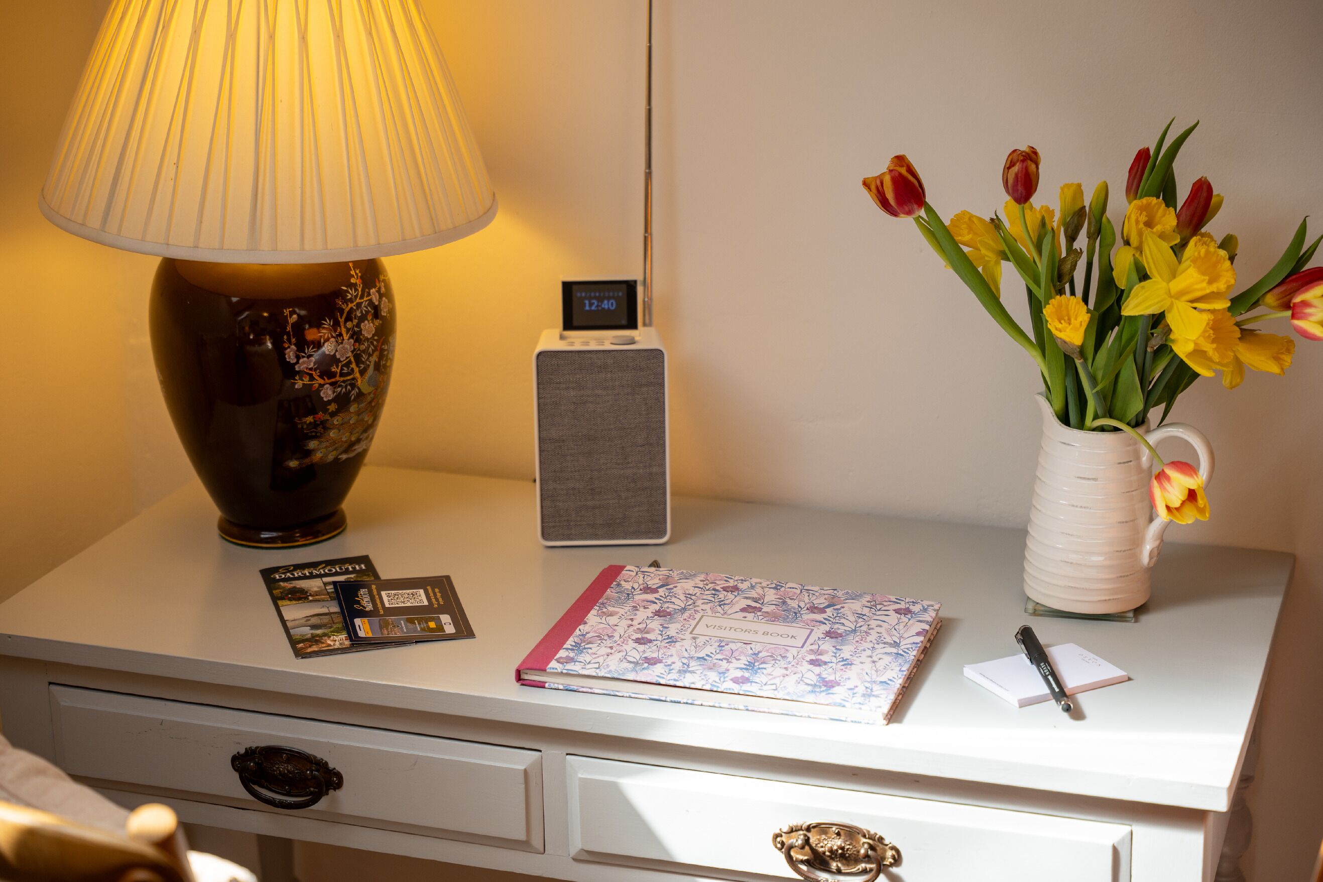 Une table de chevet avec une lampe, une horloge numérique, un livre d’or à motifs floraux pour les visiteurs, un bloc-notes et un vase contenant des tulipes jaunes et rouges.
