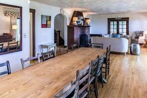 A dining area with a long wooden table, chairs, a brick fireplace, and a sofa in the background.