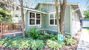 Une maison de plain-pied avec un porche à l’avant, un petit jardin avec différentes plantes et un garage attenant, situé sur le côté droit.