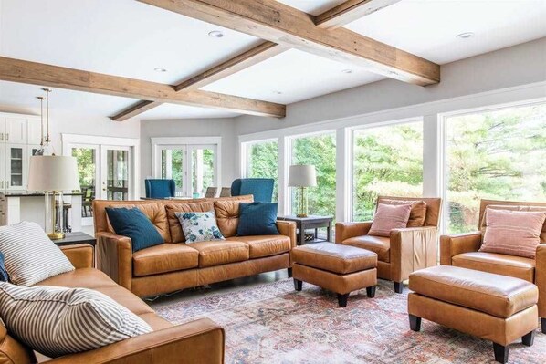 A spacious living room with leather sofas and ottomans, large windows with a view of greenery, and exposed wooden beams on the ceiling. - Dog-friendly house with private heated outdoor pool, hot tub & Ping-Pong (Grand Beach)