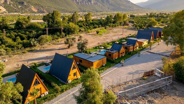 A row of wooden cabins with metal roofs, surrounded by greenery and mountains in the background.