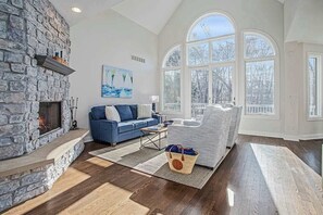 Ein Wohnzimmer mit Kamin aus Stein, einem blauen Sofa, einem grauen Sessel und großen Fenstern.