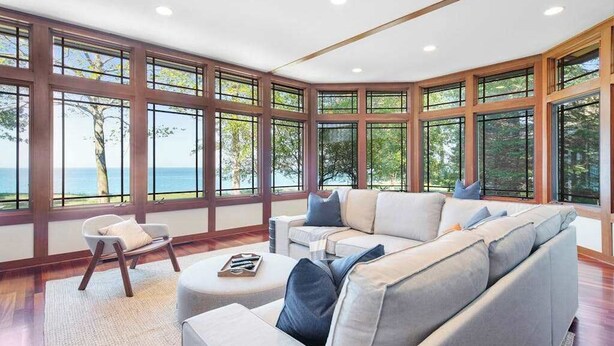 Ein modernes Wohnzimmer mit einer großen Eckcouch, einem kleinen Sessel und einem Couchtisch. Das Zimmer verfügt über große Fenster mit Blick auf das Meer und Bäume.