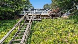 A house with a wooden staircase leading up to a deck, surrounded by lush greenery.