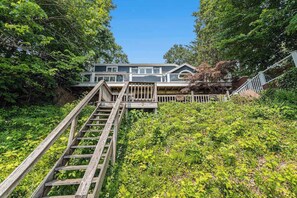 A house with a wooden staircase leading up to a deck, surrounded by lush greenery.