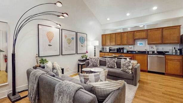 Una sala moderna con un sofá gris, una mesita de centro y una lámpara de pie. Se observa una zona de cocina con armarios de madera y un lavavajillas.
