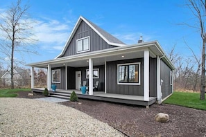 A modern two-story house with a covered porch, large windows, and a well-maintained lawn. - Modern home with large sun-room, firepit, grill, washer/dryer, & central AC (Sawyer)