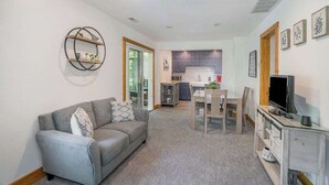 Een woonkamer met een grijze bank, een eettafel met stoelen, een televisie op een houten dressoir, en een keuken op de achtergrond.
