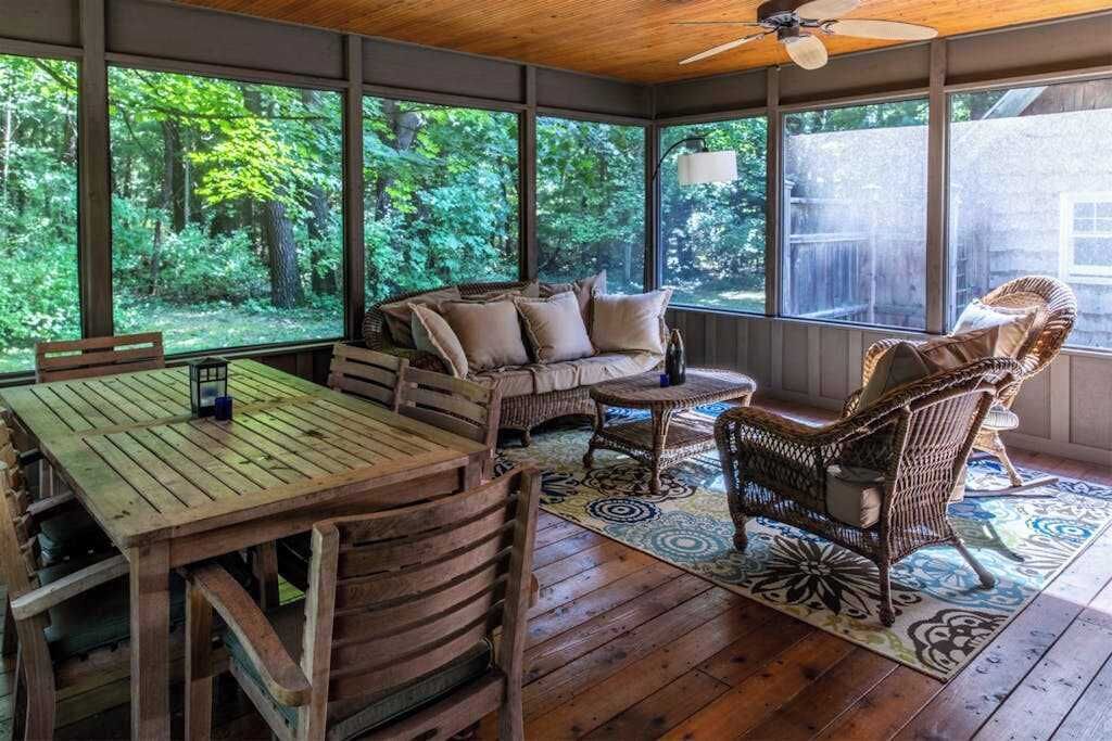 Eine Veranda mit Holzmöbeln, großen Fenstern und Blick auf einen Wald.