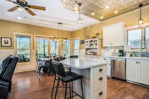 A kitchen with a central island, bar stools, a dining table, and a ceiling fan. - Dreamy retreat near the lake with spacious deck, foosball, Ping-Pong & kayaks (Saugatuck)