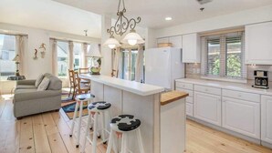 Una cocina moderna con armarios blancos, una isla central con taburetes, una zona de comedor con sofá y sillas, y una araña.