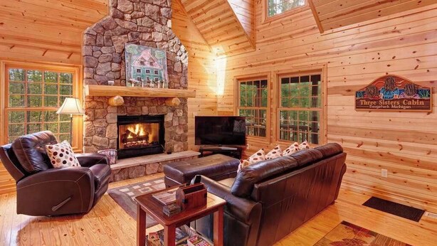 A log cabin interior with a stone fireplace, leather furniture, and a television. - Cabin tucked in the trees with private hot tub & lake/pool access (Saugatuck)