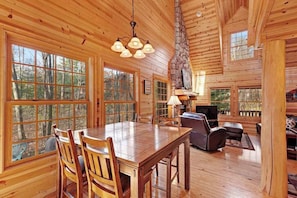 A wooden cabin interior with a dining table, chairs, a fireplace, and large windows.