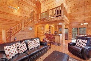 A spacious log cabin interior with a living room, dining area, kitchen, and a staircase leading to an upper level.