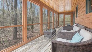 A covered porch with wicker furniture, large windows, and a view of a forest. - Cabin tucked in the trees with private hot tub & lake/pool access (Saugatuck)