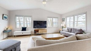 Un salón amplio con un televisor de pantalla plana grande, un ventilador de techo y vistas al exterior a través de amplias ventanas.