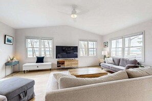 A spacious living room with a large flat-screen TV, a ceiling fan, and a view of the outdoors through large windows. - Tranquil Lakeview Retreat in Suttons Bay, with a private sauna, & large yard (Suttons Bay)