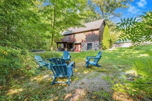 A rustic cabin with a stone foundation, a porch with chairs, and a lush green surrounding.