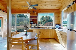 Interior de una cabaña de madera con una mesa de comedor, sillas, ventilador de techo, cocina con refrigerador y ventanas amplias que dan a un paisaje arbolado.