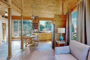 Interior de una cabaña de madera con comedor, cocina y un sofá junto a una ventana.