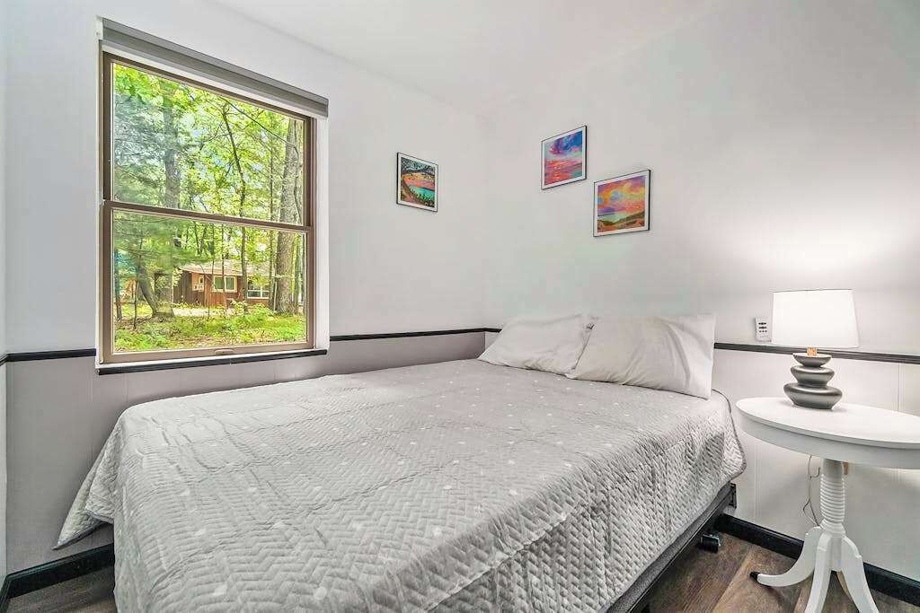 A bedroom with a large window, a bed, a bedside table with a lamp, and three framed pictures on the wall.