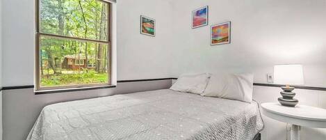 A bedroom with a large window, a bed, a bedside table with a lamp, and three framed pictures on the wall.