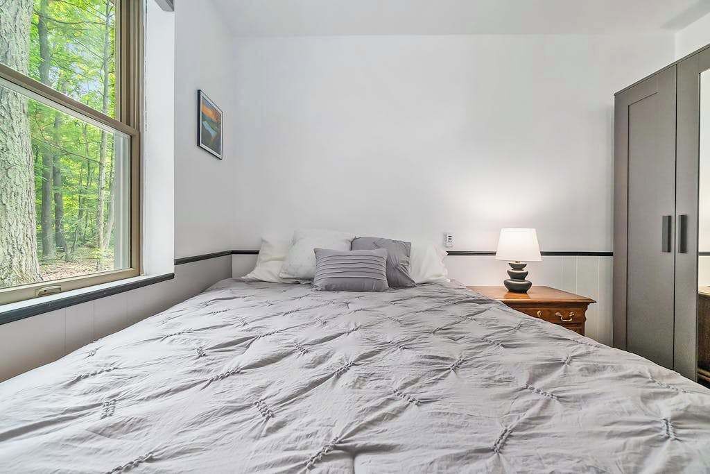 A bedroom with a bed, a nightstand, a lamp, a wardrobe, and a window with a view of trees.