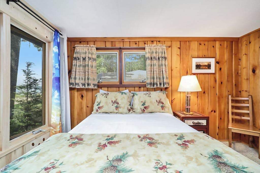A wooden-paneled room with a bed, a window with curtains, a nightstand with a lamp, and a chair.