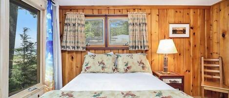 A wooden-paneled room with a bed, a window with curtains, a nightstand with a lamp, and a chair.