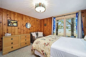 A wooden-paneled room with a bed, a dresser, a mirror, and a window with a view of trees and a body of water.