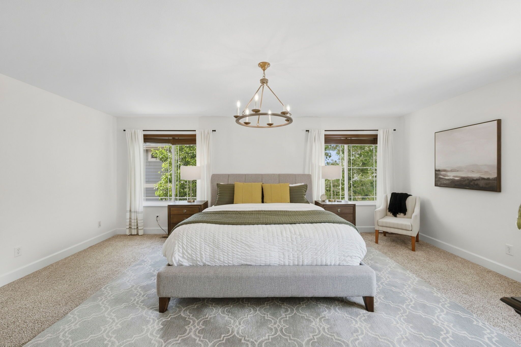 A bedroom with a large bed, two bedside tables, a chair, a chandelier, and a window with curtains.
