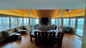 Amplia habitación con techo de madera, chimenea, una mesa de comedor con sillas y grandes ventanas con vistas.
