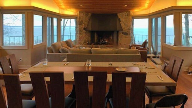 Un comedor con chimenea de piedra, techo de madera y ventanas amplias con vista al agua.