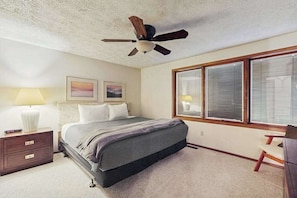 Un dormitorio con cama, mesita de noche, lámpara, silla, ventilador de techo y ventana grande.