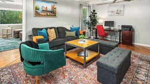 A living room with a sofa, coffee table, and ottoman, a desk with a computer, and a window with curtains. - Sandstone Hollow | King Suite, Office, Fenced Yard (Fort Collins)