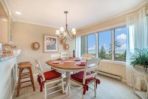 A dining area with a round table, chairs, a chandelier, and a view of the outdoors. - Lake-view condo with private beach, fireplace, deck, washer/dryer, streaming (Glen Arbor)