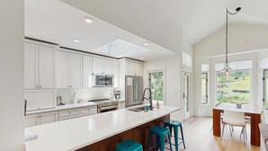 Una cocina moderna con armarios blancos, una isla grande con taburetes de bar, electrodomésticos de acero inoxidable y vistas al exterior a través de amplios ventanales.