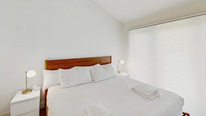 Una cama bien hecha con cabecera de madera, ropa blanca y una toalla doblada al lado.
