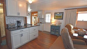 Een keuken met witte kastjes, een backsplash in visgraatmotief en een koffiezetapparaat. Er is een eethoek met een tafel en stoelen, en een raam met gordijnen.