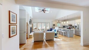 Un vaste espace de vie doté d’une grande fenêtre, d’un ventilateur de plafond et d’une cuisine avec des armoires blanches et des appareils en acier inoxydable.