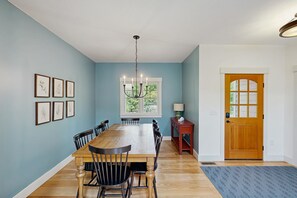 Une salle à manger avec une table et des chaises en bois, un lustre, des reproductions botaniques encadrées accrochées au mur, une fenêtre donnant sur un paysage verdoyant et une porte en bois.
