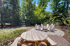 Une terrasse extérieure en bois, avec une table ronde et des chaises Adirondack blanches, entourée d’une zone boisée.