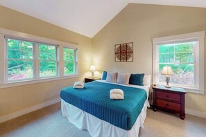 A bedroom with a large bed, two bedside tables, a wall-mounted artwork, and large windows with a view of greenery. - Downtown home near the lake with washer/dryer, firepit, backyard (Glen Arbor)
