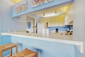 Una cocina moderna con armarios blancos, encimera blanca y pileta de acero inoxidable. Hay dos sillas de mimbre frente a la encimera.