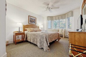Une chambre à coucher comprenant un lit, des tables de chevet, un ventilateur de plafond, une fenêtre avec des rideaux et une commode en bois.