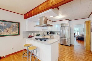 A modern kitchen with white cabinetry, a central island with stools, a stainless steel range hood, and a large map on the wall. - Dog-friendly downtown Glen Arbor home with deck, gas grill, and washer/dryer (Glen Arbor)