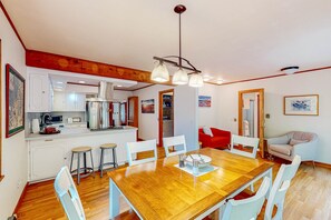 A dining area with a wooden table, white chairs, a chandelier, and a kitchen in the background. - Dog-friendly downtown Glen Arbor home with deck, gas grill, and washer/dryer (Glen Arbor)