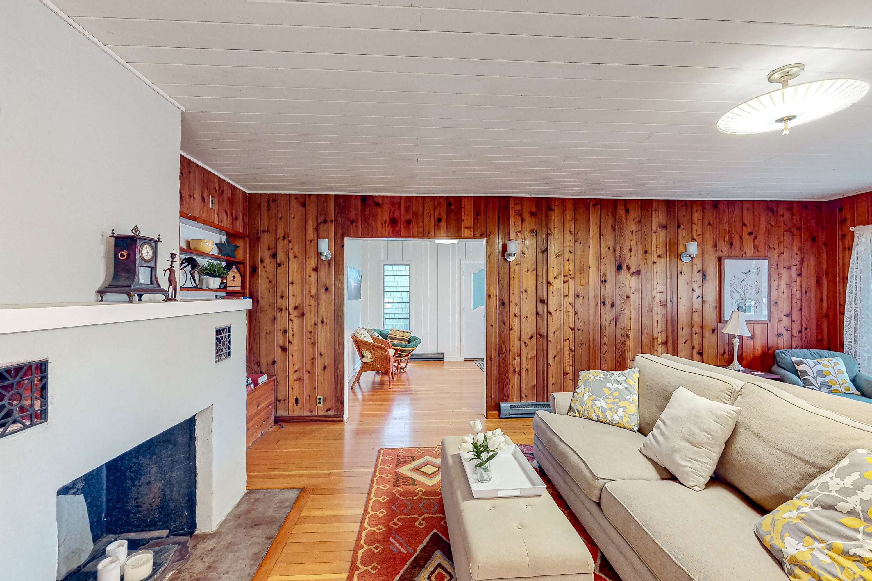 A living room with a sofa, a coffee table, a fireplace, wooden paneling, and a dining area in the background.