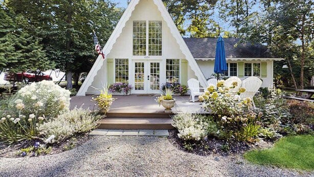 Un bâtiment blanc en forme de triangle, doté d’un porche et entouré d’un jardin où poussent diverses plantes et fleurs.