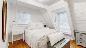 Une chambre avec un plafond en pente, un lit recouvert d’une courtepointe blanche, une commode et deux fenêtres équipées de stores.